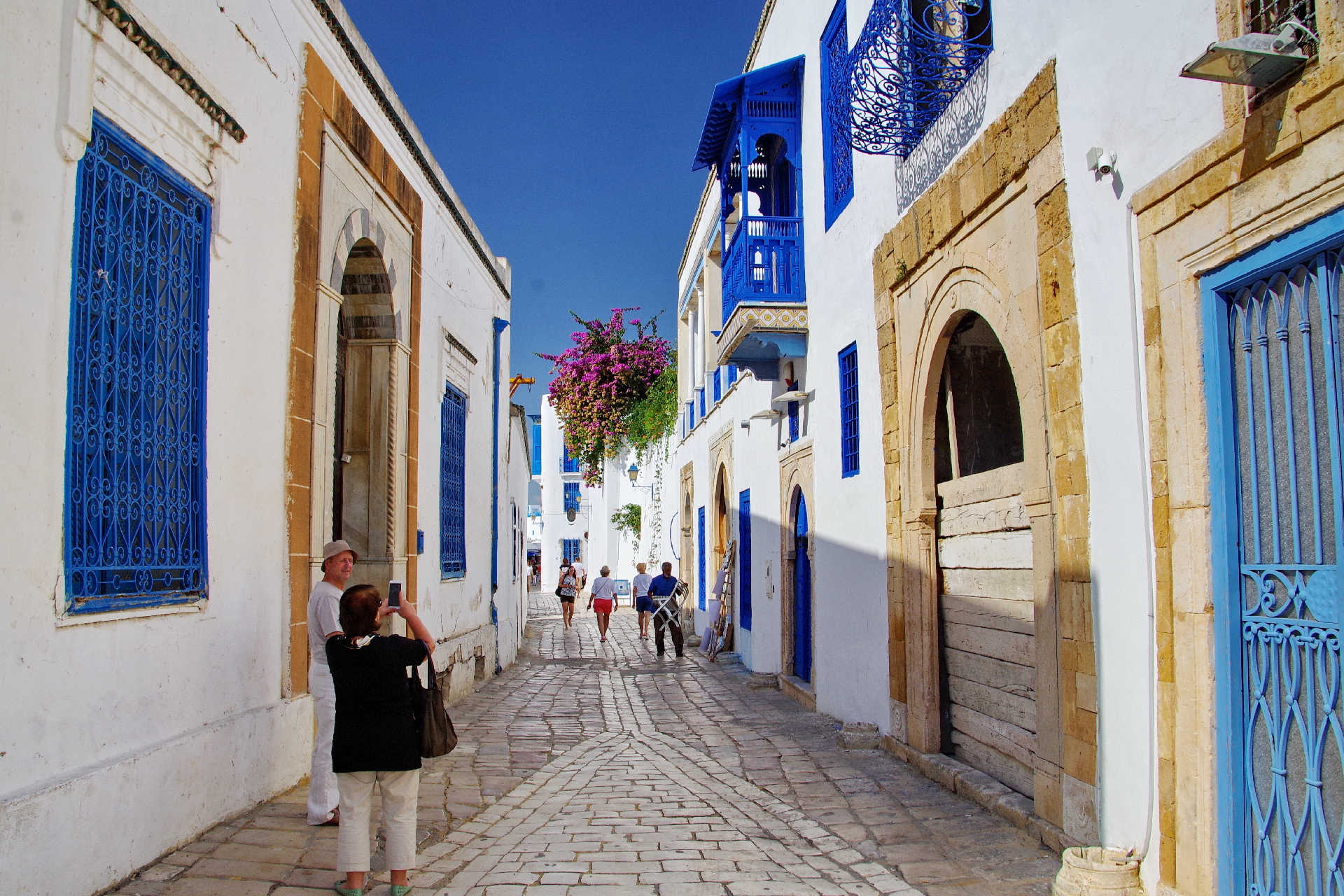 Weiß-blaue Altstadtgasse mit Bougainvillea und Touristen bei Sonnenschein