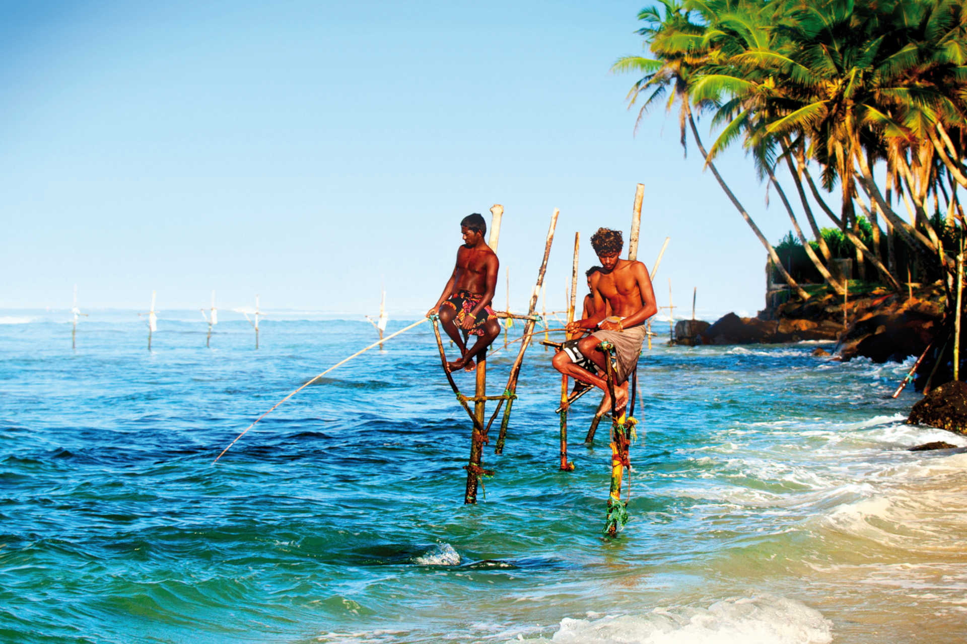 Fischer sitzen auf Holzpfählen im Meer und angeln bei Wellen und Palmen am Ufer