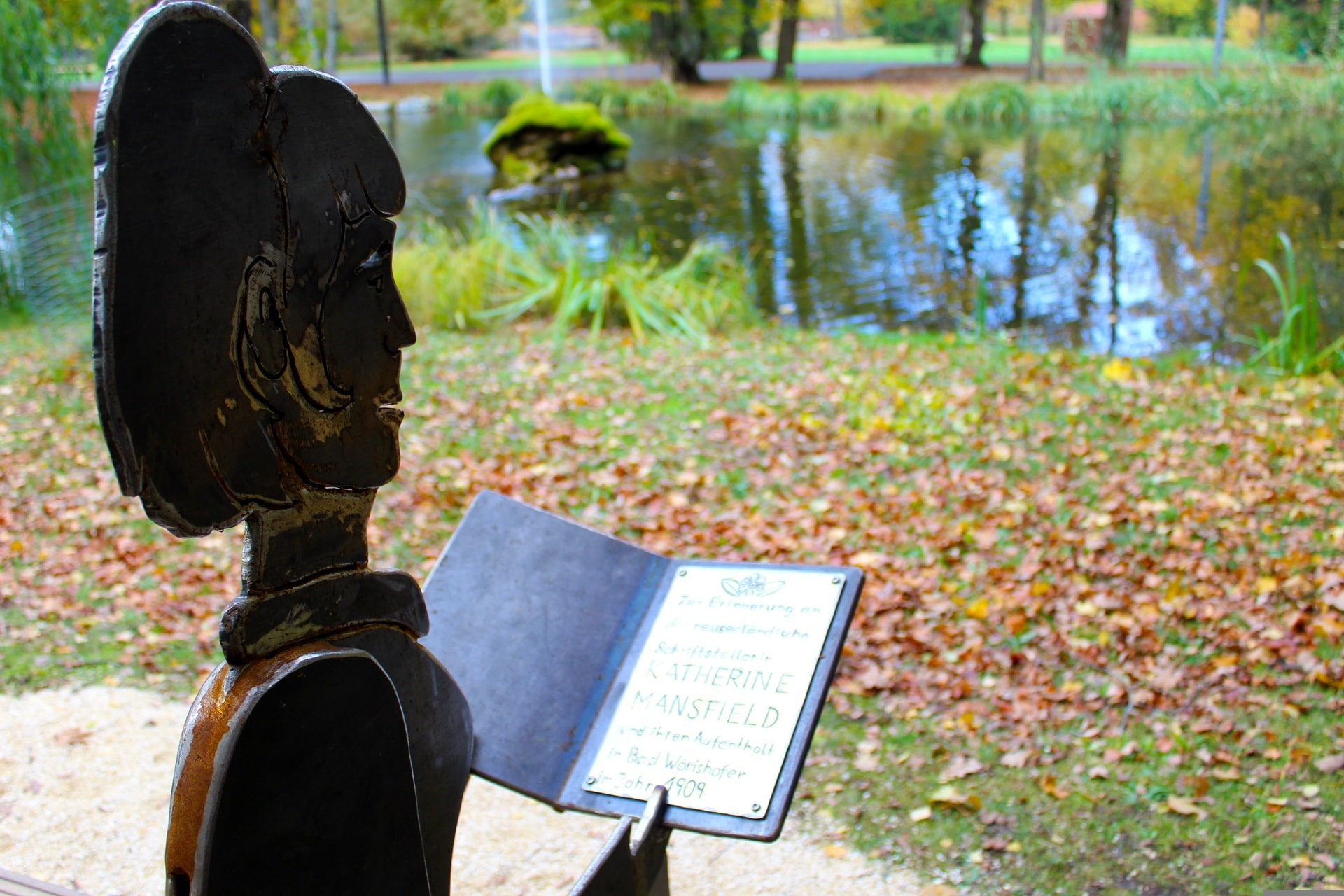Metallfigur mit Buch im Park vor Teich und herbstlichem Laub