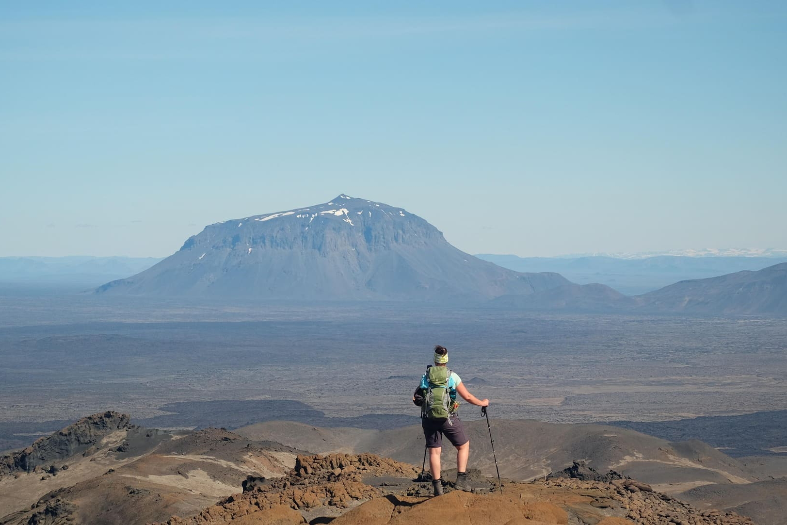 Person mit Rucksack blickt über weite Lavalandschaft zum Berg