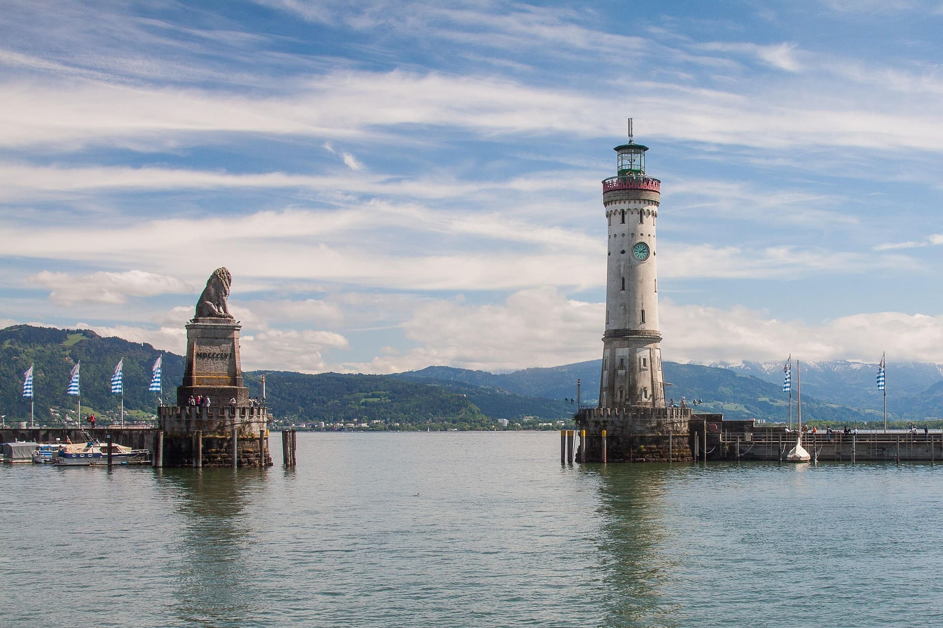 Löwenstatue und weißer Leuchtturm am Bodensee vor Alpenkulisse