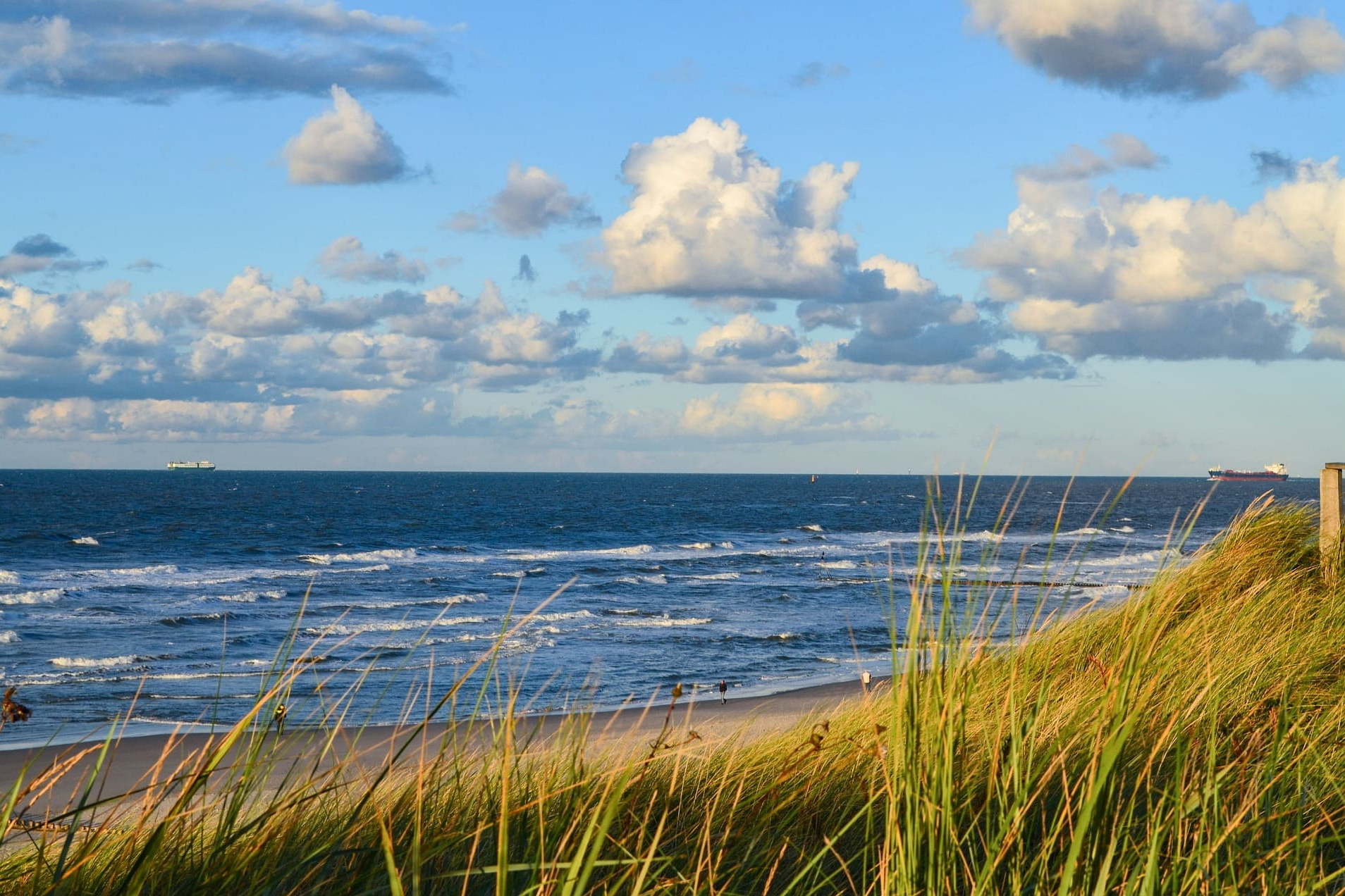 Wellen am Sandstrand hinter Dünen mit hohem Gras und Wolkenhimmel