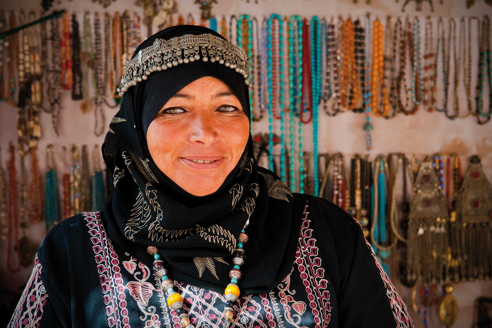 Frau in traditioneller Kleidung mit Kopftuch und buntem Schmuck vor Marktstand