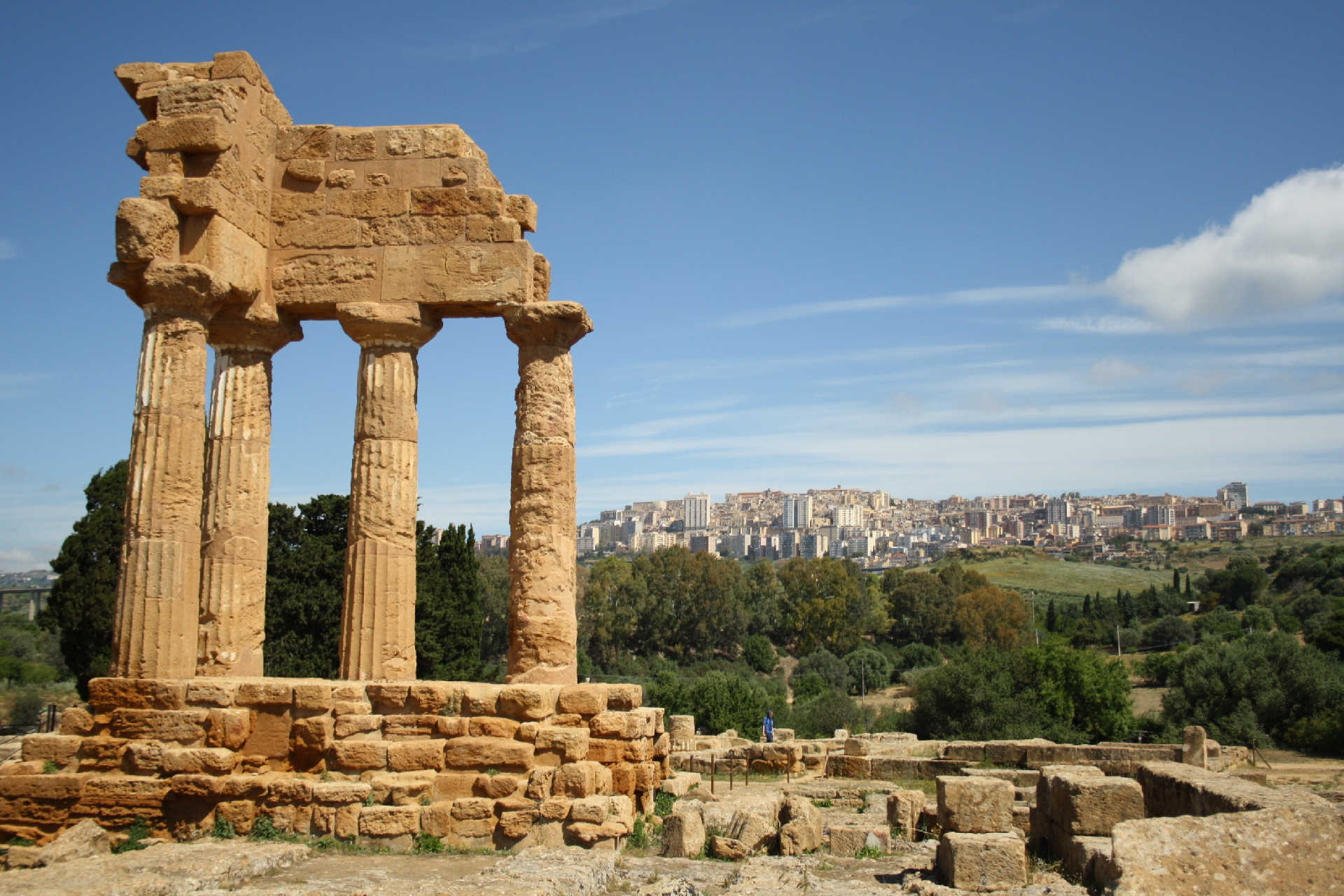Antike Tempelreste mit vier dorischen Säulen im Tal der Tempel bei Agrigent auf Sizilien, im Hintergrund die moderne Stadt auf einem Hügel.