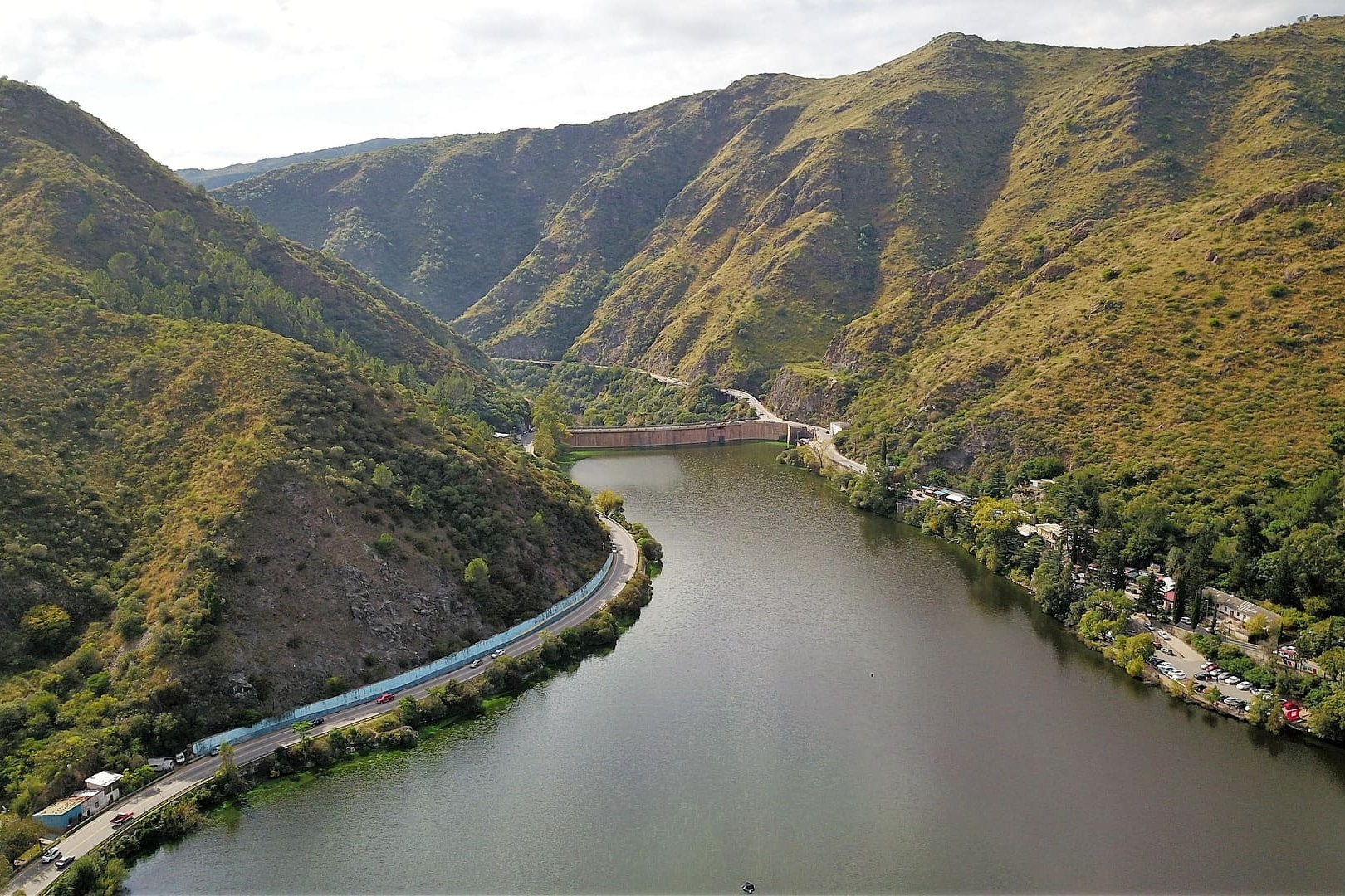 Stausee inmitten grüner Berge mit Straße am Ufer und Blick auf die Staumauer