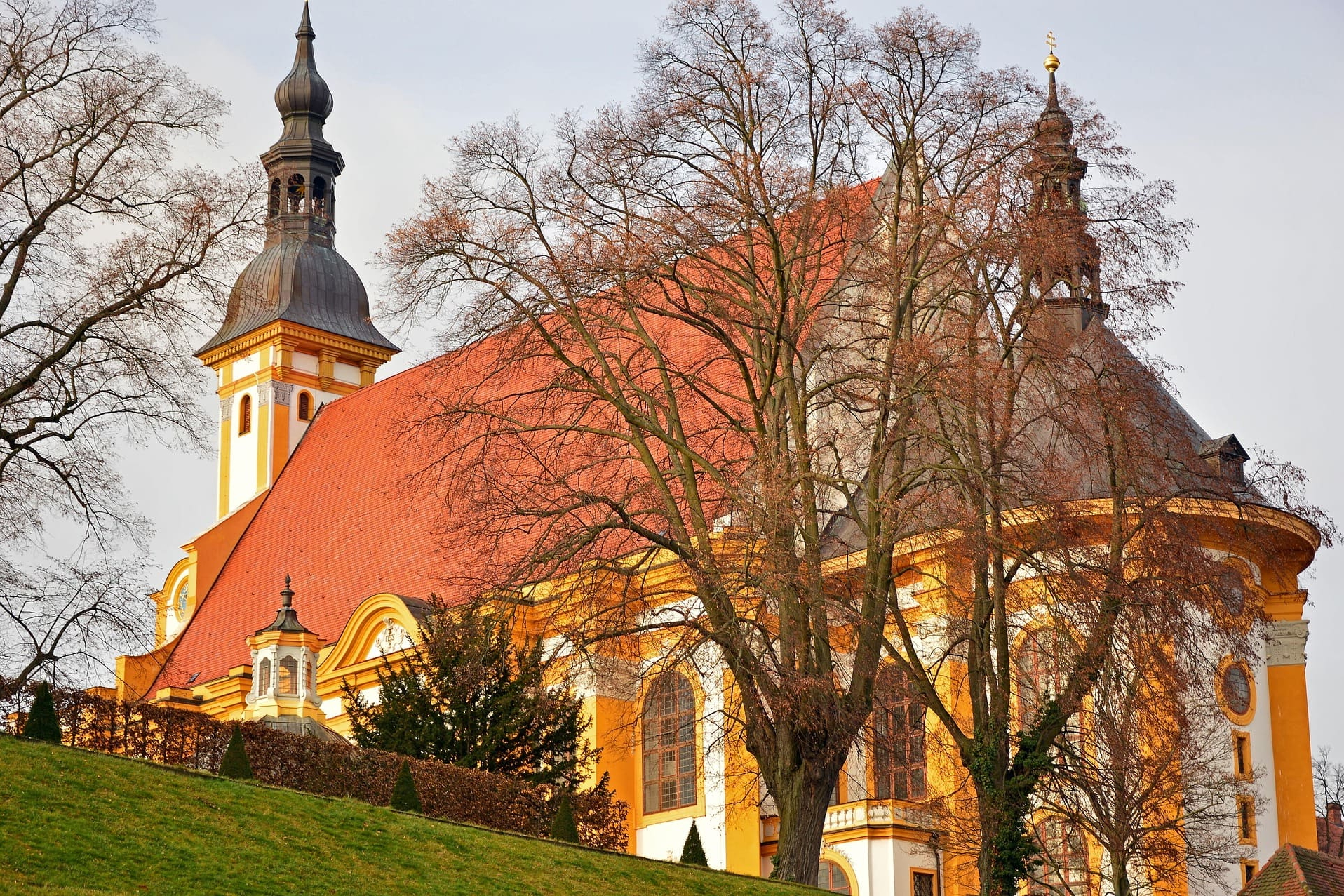 Gelbweiße Barockkirche mit rotem Dach hinter kahlen Bäumen im Winter