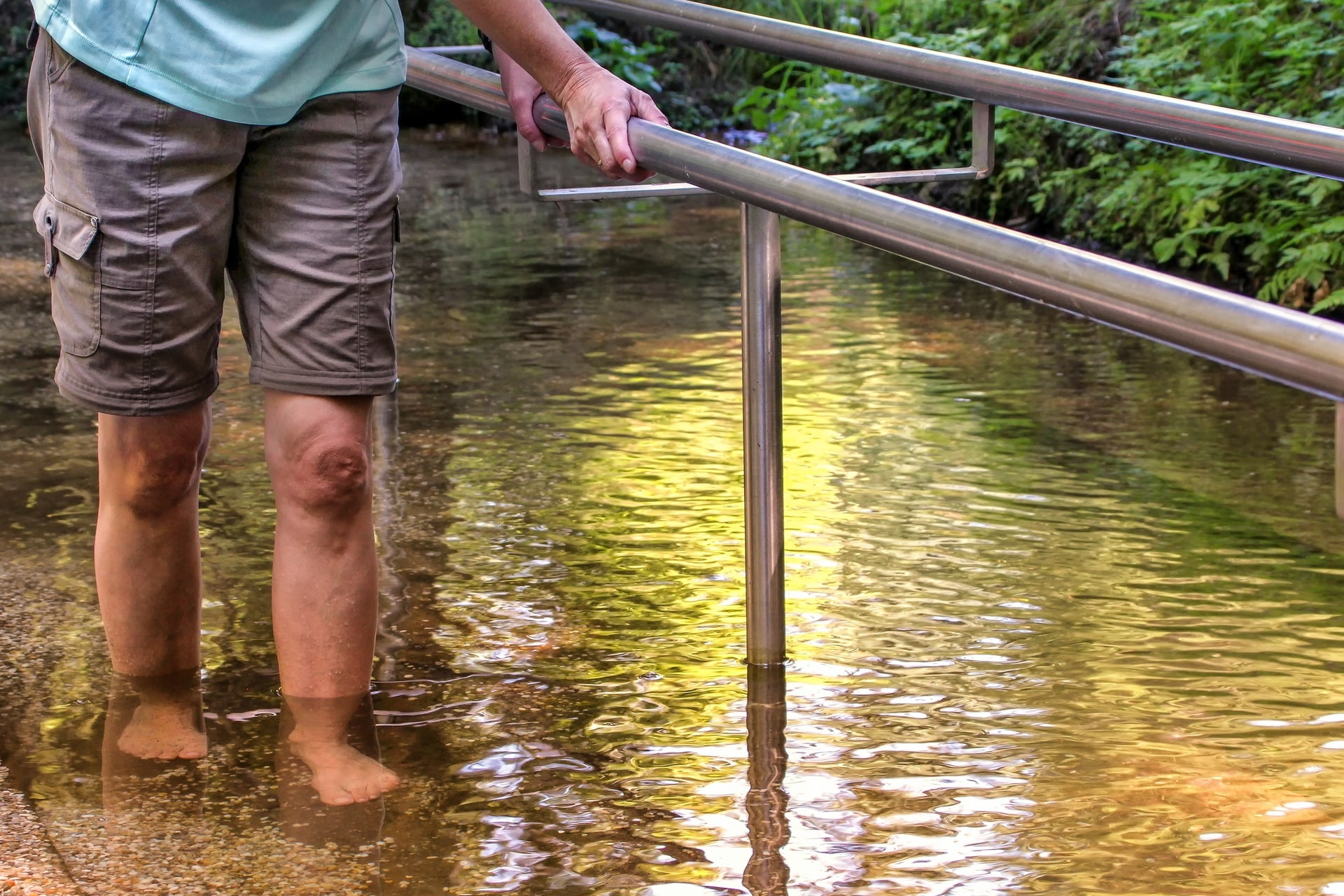 Person geht barfuß im flachen Wasser entlang eines Metallgeländers