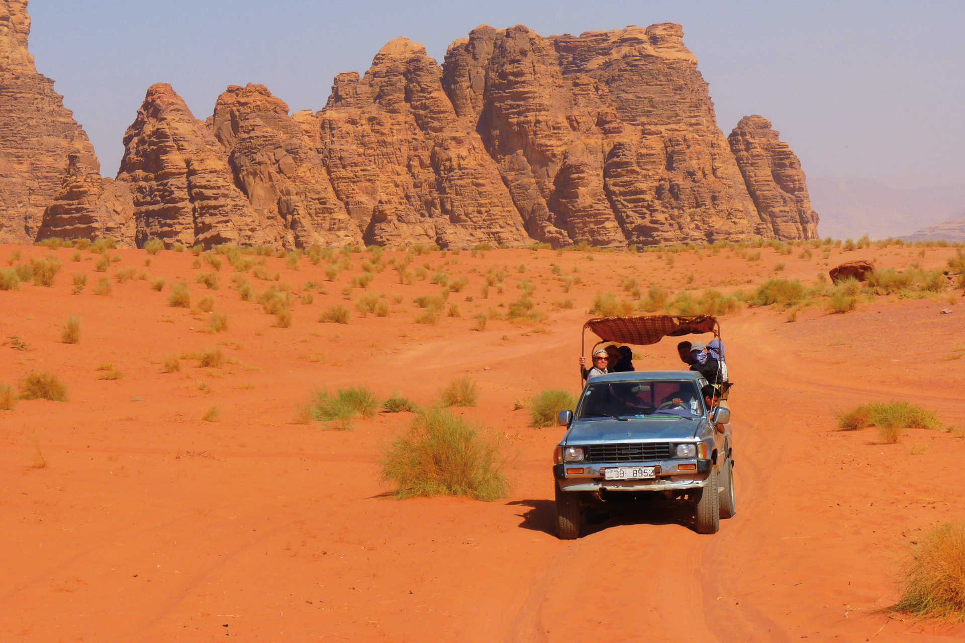 Geländewagen mit Touristen fährt durch rote Wüste vor hohen Felsformationen