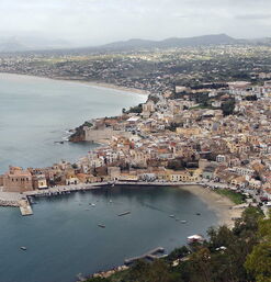 Blick auf die sizilianische Küste mit der historischen Altstadt von Castellammare del Golfo und der markanten Festung am Hafen, eingerahmt von sanften Hügeln und dem weiten Mittelmeer.