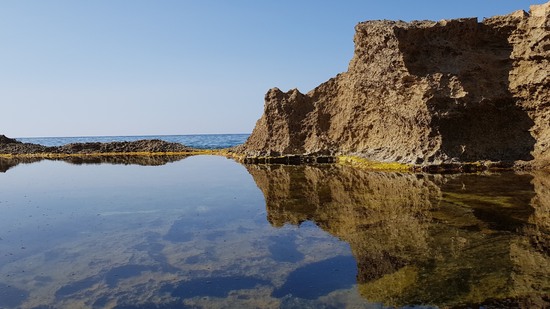 Klares Wasserbecken spiegelt Felsen vor blauem Himmel am Meer