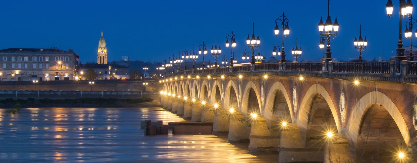 Beleuchtete Steinbrücke mit Laternen über Fluss bei Nacht in Bordeaux